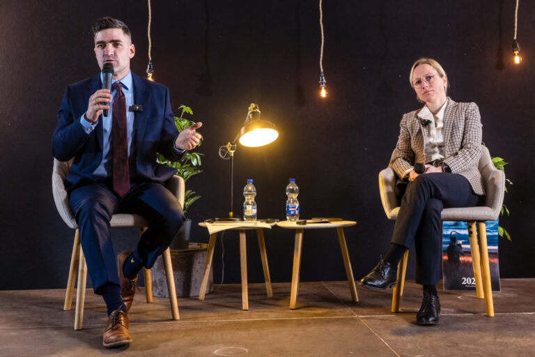 Martin Sellner und Lena Kotré sitzen auf einer Bühne auf Stühlen und halten jeweils ein Mikrofon, zwischen ihnen stehen kleine Tische mit Wasserflaschen und eine Lampe vor dunklem Hintergrund. Auftritt von Martin Sellner bei einer Veranstaltung im Spreewald, an der auch die Brandenburger AfD-Landtagsabgeordnete Lena Kotré trotz vorheriger Intervention der Parteispitze teilnahm. Martin Sellner neben Brandenburgs AfD-Abegordneten Lena Kotré: Diskussion über Remigration. Foto: picture alliance/dpa | Frank Hammerschmidt