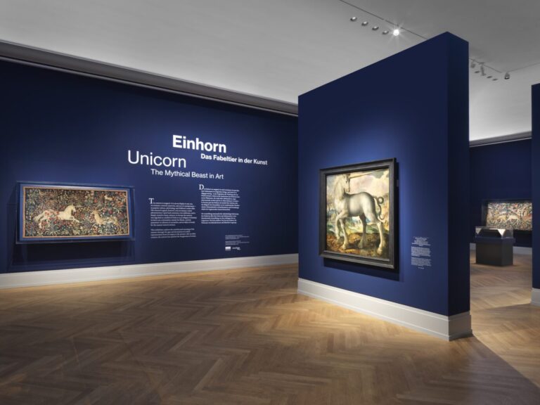 In einem stimmungsvoll beleuchteten Raum sieht man blaue Wände, an diesen hängen Gemälde, die Einhörner zeigen – es handelt sich um einen Raum der Ausstellung „Einhorn. Das Fabeltier in der Kunst“ des Museum Barberini in Potsdam