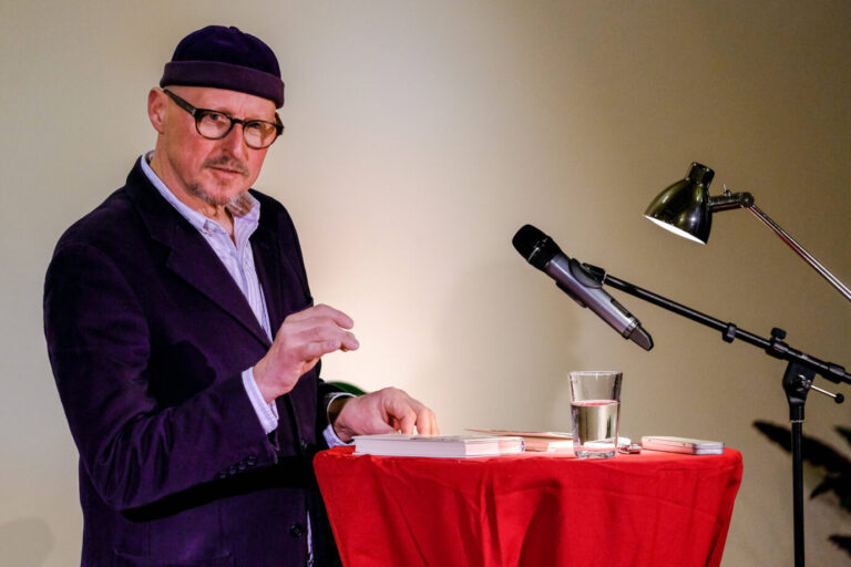 Mit schwarzer Mütze und in schwarzer Jacke steht der Schriftsteller und Lyriker Jörg Bernig vor einem kleinen Tisch.