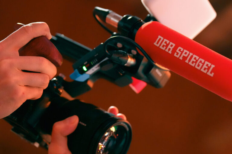 Eine Person hält ein Mikrofon mit der Aufschrift „DER SPIEGEL“ während einer Aufnahme oder eines Interviews. Im Vordergrund ist eine Kamera mit großem Objektiv zu sehen. Fotograf im Einsatz für den „Spiegel“: Das Magazin entfernte KI-manipulierte Bilder einer Agentur. Foto: picture alliance / Flashpic | Jens Krick