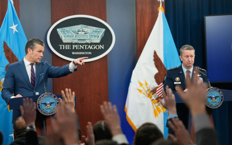 Pete Hegseth spricht am Rednerpult im Pentagon und zeigt mit dem Finger ins Publikum, neben ihm steht Generalstabschef Dan Caine; im Vordergrund heben Journalisten die Hände. Kontext: Pressekonferenz zu den US-Militäroperationen gegen den Iran in Washington. Pete Hegseth bei der Pressekonferenz im Pentagon: Der US-Verteidigungsminister verteidigt die Angriffe auf den Iran und kündigt weitere Schritte an. Foto: picture alliance / ASSOCIATED PRESS | Mark Schiefelbein