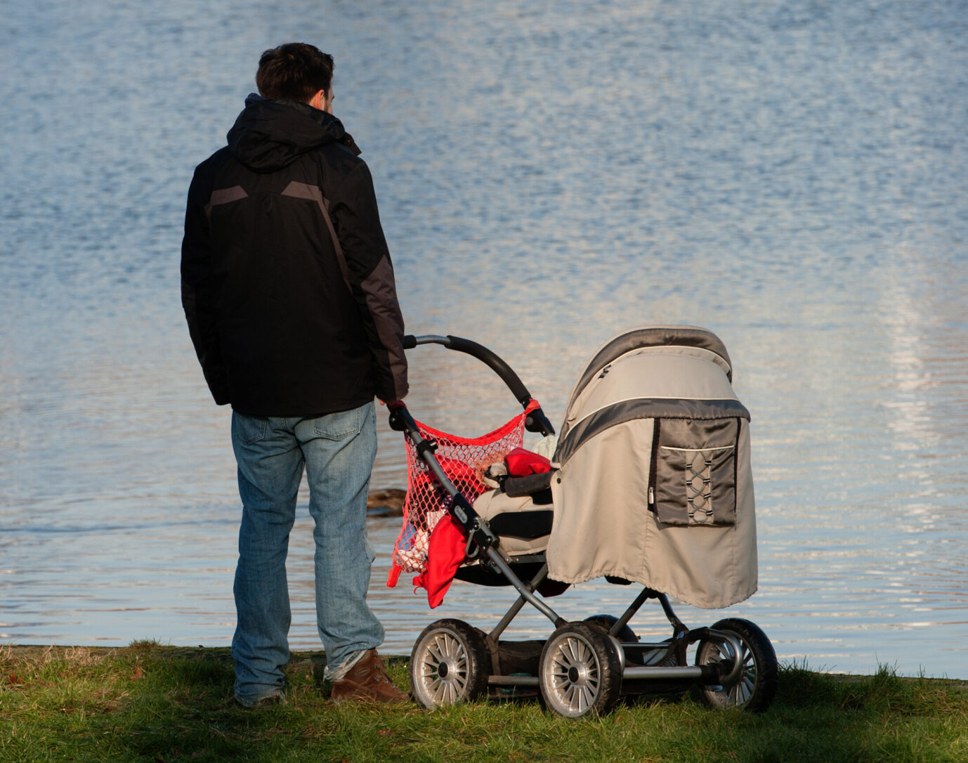 Ein Mann steht mit einem Kinderwagen am Ufer eines Sees und blickt auf das Wasser. Symbolbild für einen alleinerziehenden Vater, dem nach einem Internetkommentar mit der Parole „Alles für Deutschland“ eine Vorstrafe und der Verlust seines Arbeitsplatzes drohen. Alleinerziehender Vater mit Kinderwagen am See (Symbolbild): Ein Strafbefehl wegen der Parole „Alles für Deutschland“ könnte einen Mann seinen Arbeitsplatz kosten. Foto: picture alliance / dpa | Paul Zinken