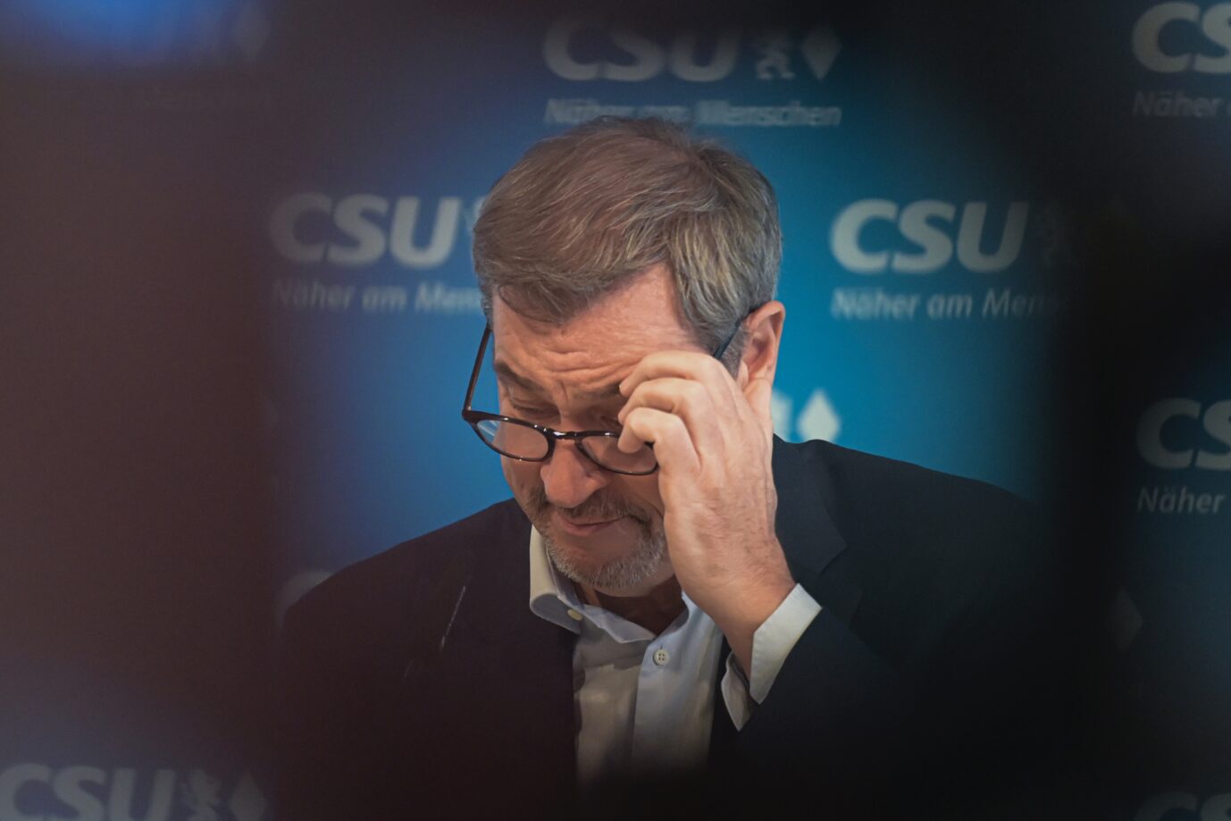 Ein Mann mit Brille und Anzug reibt sich mit der Hand über die Stirn, im Hintergrund ist das CSU-Logo mit dem Slogan „Näher am Menschen“ zu sehen. Der bayerische Ministerpräsident wirkt nachdenklich während einer Veranstaltung. Markus Söder bei einer Pressekonferenz der CSU: das Wahlergebnis scheint ihm zu schaffen zu machen. Foto: picture alliance/dpa | Malin Wunderlich