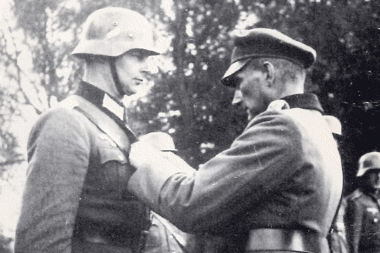 Schwarzweißfotografie: Zwei Männer in militärischer Uniform stehen einander gegenüber, einer mit Stahlhelm, der andere mit Schirmmütze, und befestigen etwas an der Brust des Soldaten; im Hintergrund sind unscharf weitere uniformierte Personen und Bäume zu erkennen. Oberleutnant Prinz Wilhelm von Preußen wird im Oktober 1939 mit dem Eisernen Kreuz zweiter Klasse ausgezeichnet: Er habe fest damit gerechnet zu fallen – ohne jemals die Krone getragen zu haben. Foto: BeBra Verlag