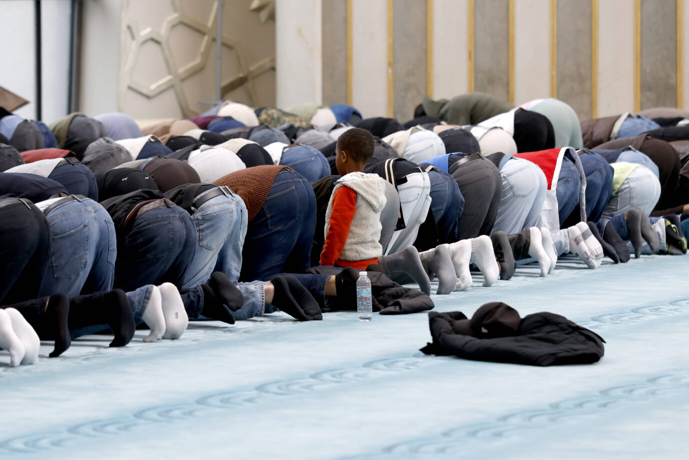Innenansicht der DITIB Zentralmoschee in Ehrenfeld am Tag der offenen Moschee . An der Kölner Zentralmoschee darf seit 2022 freitags per Lautsprecher von einem Muezzin zum Gebet gerufen werden. Mehrere Reihen von Männern knien dicht nebeneinander auf einem Teppichboden während eines islamischen Gebets; in der Mitte sitzt ein Kind aufrecht zwischen den Betenden, vor ihm liegen eine Wasserflasche und eine Jacke. Gemeinschaftsgebet von Muslimen in einer Moschee. Moslems beim Gebet in einer Moschee: Laut Wahltagsbefragungen wählen viele von ihnen links. Foto: IMAGO / Panama Pictures