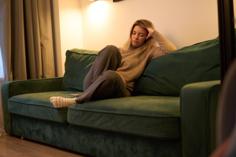 Eine junge Frau sitzt auf einem Sofa in einer Wohnung, lehnt sich zurück und stützt den Kopf mit der Hand, die Augen geschlossen. Sie wirkt erschöpft und angespannt. Im Hintergrund sind unscharf Möbel und Wohnraum zu erkennen. Das Bild steht im Zusammenhang mit einer Studie zur Jugend in Deutschland, die zunehmende psychische Belastungen, Depressionen und Zukunftssorgen bei jungen Menschen beschreibt.