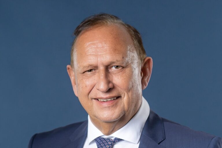 Porträt eines älteren Mannes mit kurzem, seitlich gekämmtem Haar in dunkelblauem Anzug, weißem Hemd und gemusterter Krawatte. Er blickt leicht seitlich in die Kamera und lächelt. Im Jackett steckt ein weißes Einstecktuch. Der Hintergrund ist einfarbig blau. Bernhard Pepperl im Porträt: Der AfD-Politiker gewann in Mannheim ein Direktmandat – auch gewählt von Migranten. Foto: Bernhard Pepperl