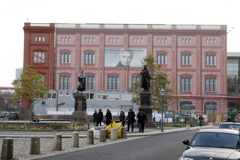 Man sieht ein rotes Gebäude und davor Statuen: Es handelt sich um Karl Friedrich Schinkels Bauakademie in Berlin, davor ein Denkmal des Architekten.