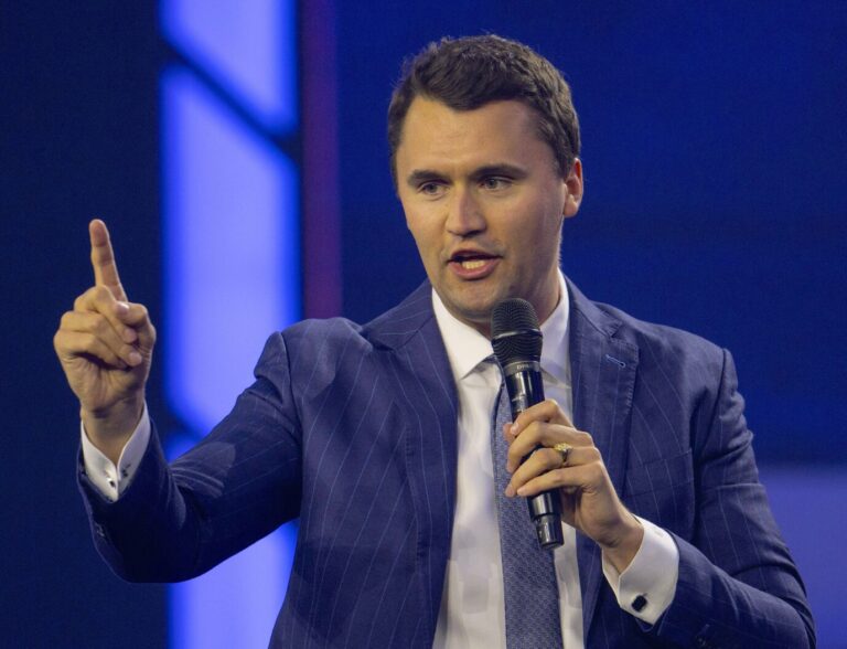 Charlie Kirk trägt Anzug und Krawatte und streckt einen Finger in die Luft.