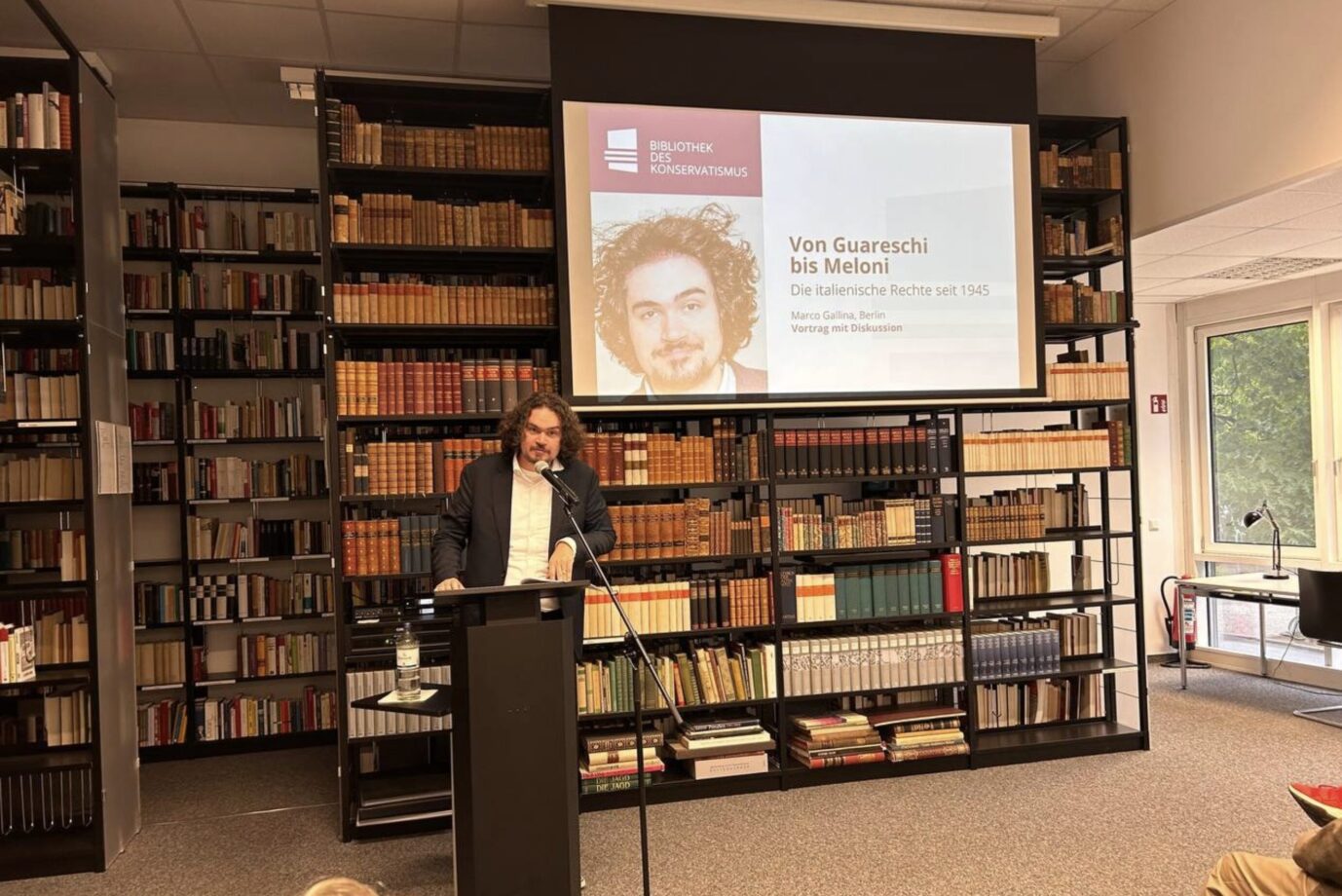 An einem Rednerpult steht der Historiker, Journalist und Politikwissenschaftler Marco Gallina und referiert über die Nachkriegsrechte Italiens – hinter ihm stehen Bücher