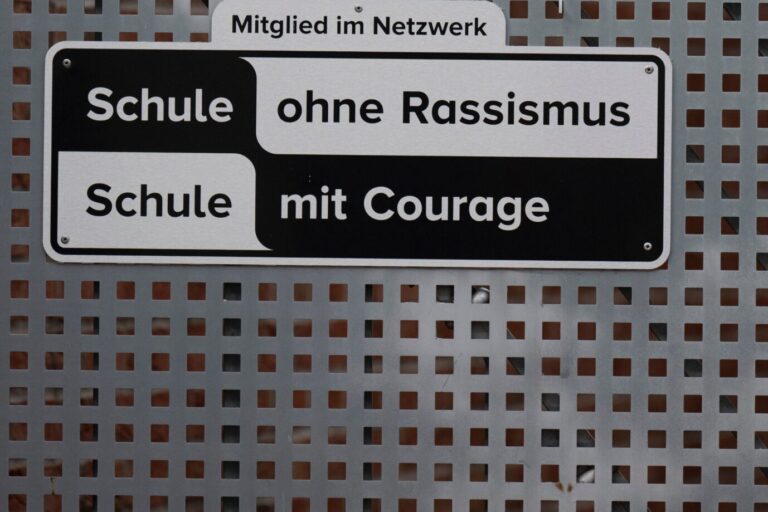 Das Bild zeigt ein Schild mit der Aufschrift „Schule ohne Rassismus – Schule mit Courage“. Das Nicolaus-Cusanus-Gymnasium in Bonn ist Teil des Netzwerks, während viele Schüler unter aggressiven Migranten leiden.