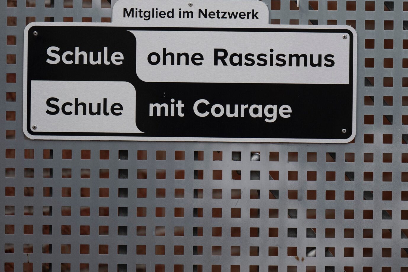 Das Bild zeigt ein Schild mit der Aufschrift „Schule ohne Rassismus – Schule mit Courage“. Das Nicolaus-Cusanus-Gymnasium in Bonn ist Teil des Netzwerks, während viele Schüler unter aggressiven Migranten leiden.
