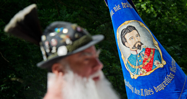 Ein bärtiger Mann mit Gamsbart steht am 15.06.2014 in Berg (Bayern) vor einem Banner mit dem Konterfei von König Ludwig II. Die "Königstreuen" kamen an der Votivkapelle im Schlosspark bei Berg zu einer Gedenkfeier anlässlich des 128. Todestages von König Ludwig II. zusammen. Foto: Nicolas Armer/dpa ++