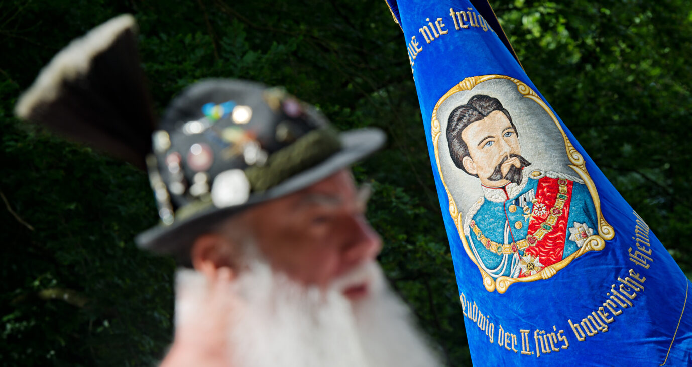 Ein bärtiger Mann mit Gamsbart steht am 15.06.2014 in Berg (Bayern) vor einem Banner mit dem Konterfei von König Ludwig II. Die "Königstreuen" kamen an der Votivkapelle im Schlosspark bei Berg zu einer Gedenkfeier anlässlich des 128. Todestages von König Ludwig II. zusammen. Foto: Nicolas Armer/dpa ++