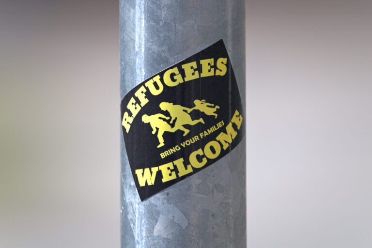 Schleswig, ein kleiner Aufkleber mit der Aufschrift: "Refugees Welcome, Bring Your Families" auf einem Verkehrsschildmast. Das Logo mit dem Piktogramm einer flüchtenden Familie wird international genutzt. Ursprünglich war das Bild ein Hinweisschild auf Highways in den USA. Es warnte Autofahrer vor illegalen Immigranten, die möglicherweise überraschend die Straße überqueren. Das finale Logo wurde vom Kollektiv um den Online-Shop "Red Stuff" erstellt. Ein „Refugees Welcome“-Aufkleber (Symbolbild): Welche Kosten eine geduldete Person Köln verursache, sei laut der Stadt technisch nicht gesondert auszuwerten. Foto: picture alliance / SULUPRESS.DE | Torsten Sukrow/SULUPRESS.DE