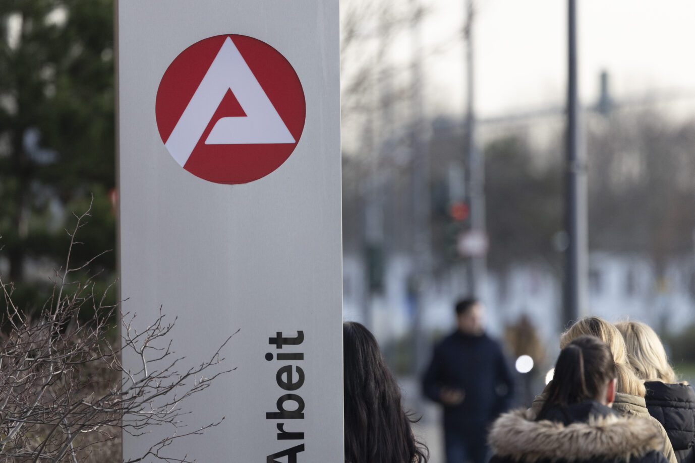 Das Logo der Agentur für Arbeit prangt auf einem Metallschild, davor laufen junge Menschen – es geht um Arbeitslosigkeit und Bildung