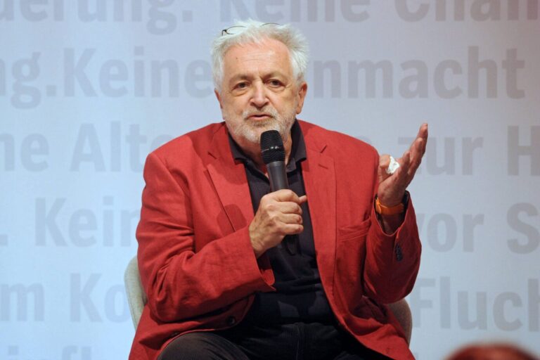 Henryk M. Broder trägt ein rotes Jackett und gestikuliert mit den Händen