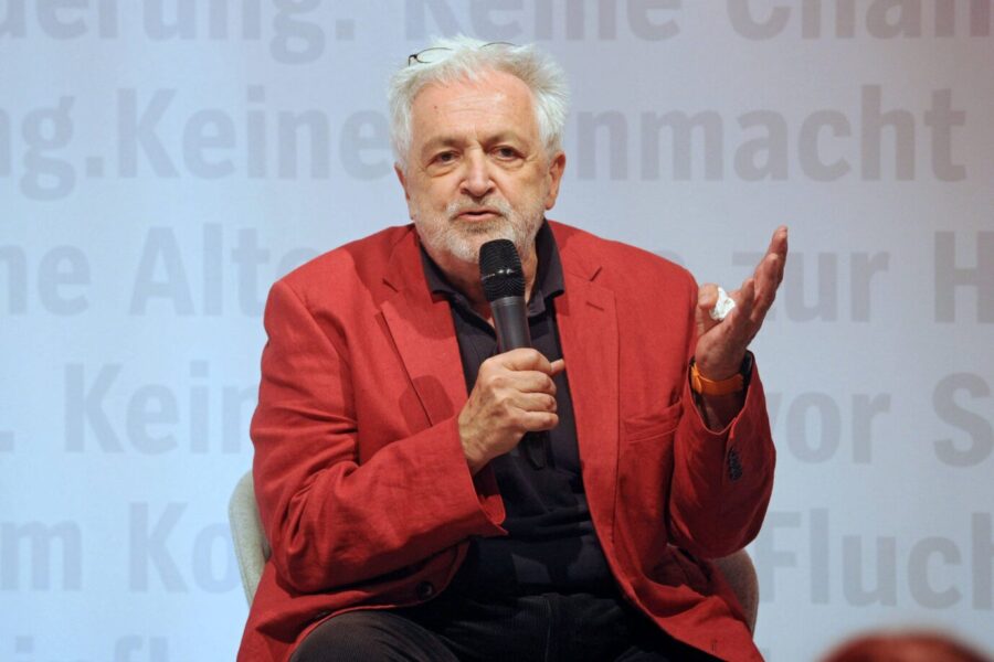 Henryk M. Broder trägt ein rotes Jackett und gestikuliert mit den Händen