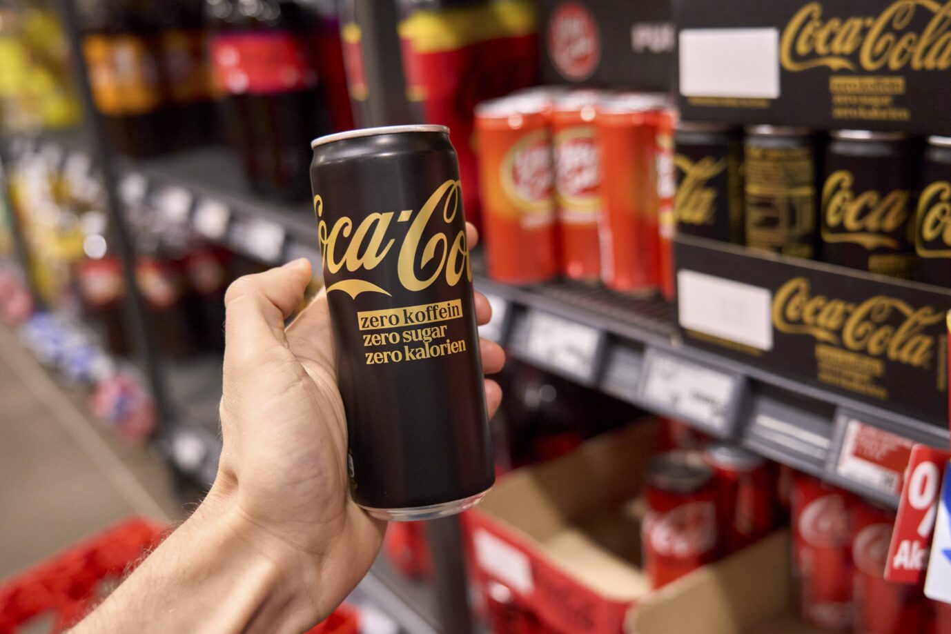 Eine Hand hält eine Dose Coca-Cola Zero in einem Supermarkt hoch – obwohl sie keinen Zucker enthält, wird sie bald teurer